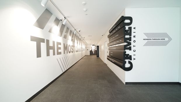 A bright, modern commercial corridor or reception passage with white graphic walls, large black typographic signage, directional arrows, and a dark grey tiled floor. Track lighting illuminates the elongated space, while a dark wall-mounted sculptural sign and photographic display anchor the far end.