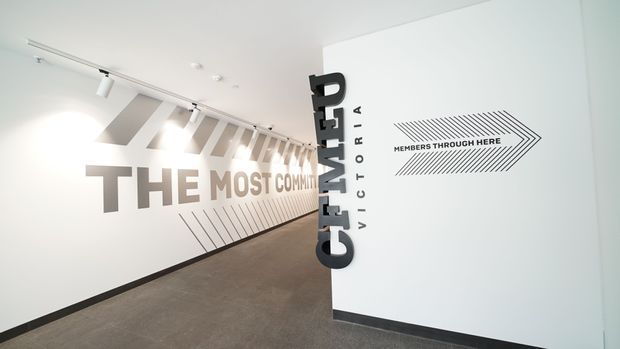 A bright, minimalist commercial interior, likely an entrance or hallway, with bold typographic wall graphics, directional arrow signage, track lighting, and a vertical “CIMET”/“Victoria” style branded feature. The space has white walls, a dark neutral carpeted floor, and strong geometric graphic elements.