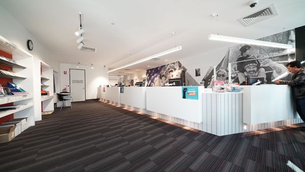 A bright contemporary commercial interior, likely a retail or reception space, with long white service counters, open display shelving, dark striped carpet, linear pendant lighting, and a large black-and-white graphic mural. The space has a clean, functional fit-out with strong graphic accents and visible customer-service activity.