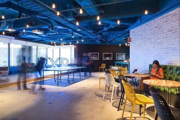 An open-plan commercial interior with an industrial-inspired café or collaborative lounge atmosphere. The space features a polished concrete floor, exposed blue-painted ceiling structure and services, suspended pendant bulbs, exposed light brickwork, extensive glazing, framed artwork, café tables, and varied colourful chairs. A ping-pong table and blurred figures suggest a flexible workplace or social recreation area.