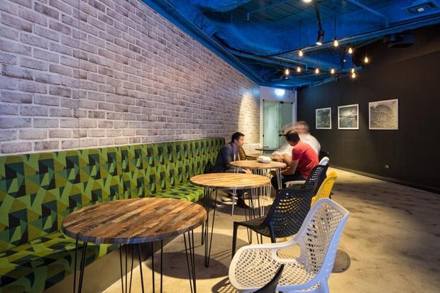 A narrow industrial-style commercial interior, likely a café or casual dining venue, with exposed painted services and ceiling, a long pale brick feature wall, dark painted walls, polished concrete flooring, and a colourful geometric upholstered bench. Round timber tables with slender black metal legs are paired with woven and perforated chairs. Warm pendant and string lighting contrasts with the blue-painted ceiling, while framed artwork decorates the rear wall.