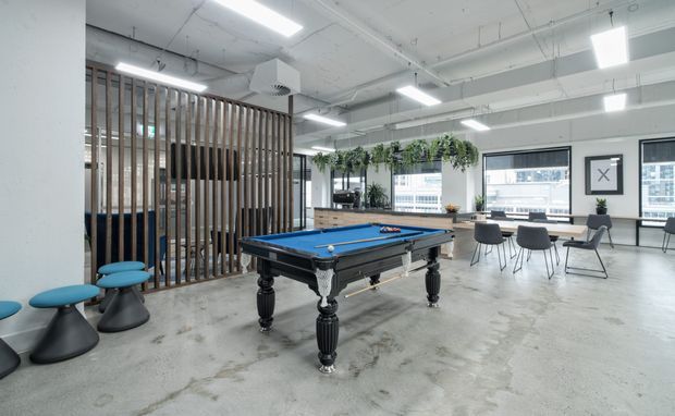 Open-plan contemporary commercial recreation and breakout space with a traditional-style blue billiards table in the foreground, casual stools to the left, a dining or meeting table by large windows, suspended greenery, and a timber slatted divider. The space has exposed concrete flooring, a white exposed-services ceiling, and bright linear and rectangular fluorescent lighting.