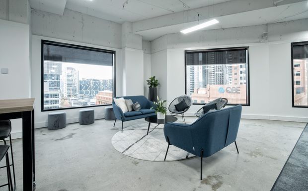 Bright contemporary office lounge with exposed concrete flooring and ceiling, white structural columns, large urban-facing windows, and a small seating arrangement of blue lounge chairs around a black round coffee table. A partial kitchenette or bar counter is visible at left, with city buildings beyond the glazing.