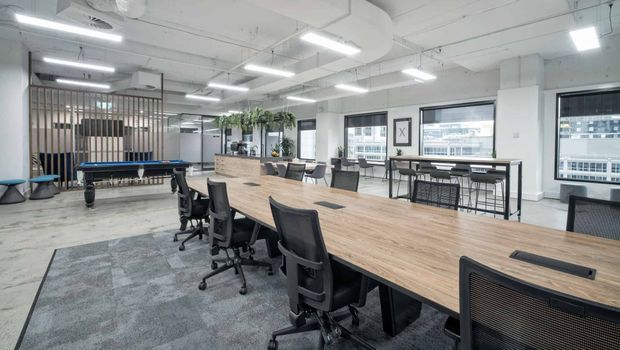 Open-plan contemporary commercial office with a long communal timber worktable, numerous ergonomic office chairs, high tables, stools, and a pool table. The bright white fit-out includes exposed ceiling services, linear fluorescent lighting, large windows, carpeted meeting/work areas, and a timber-slatted partition with indoor greenery.