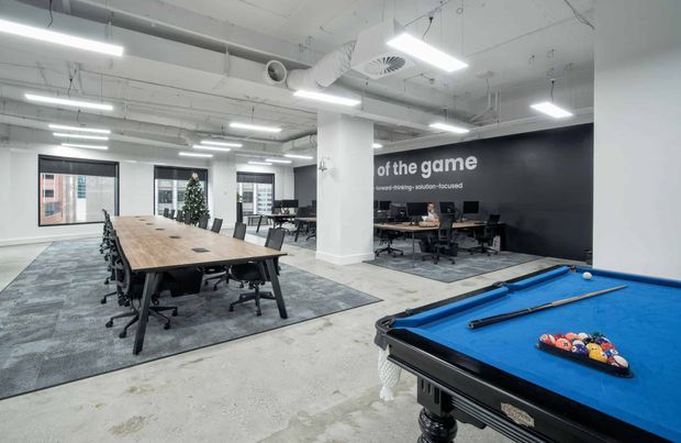 Open-plan contemporary office and recreation area with exposed services, white structural columns, linear fluorescent lighting, large communal timber meeting table, workstations, carpet zones, and a blue-felt billiards table in the foreground. A dark feature wall with motivational text defines the office backdrop.