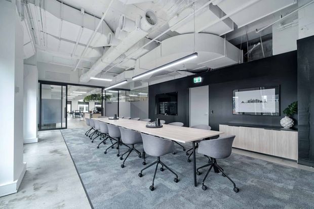 Contemporary commercial meeting room with a long light-wood conference table surrounded by grey upholstered swivel chairs. The space features dark charcoal feature walls, light wood built-in cabinetry, glass-partitioned offices, exposed white services and ductwork, linear pendant lighting, and grey carpet over a polished concrete floor.