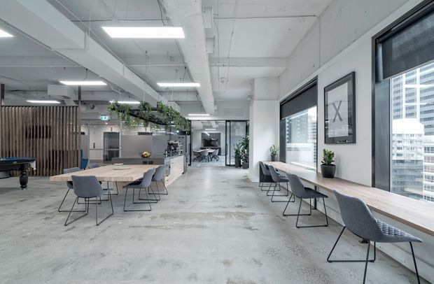A bright, open-plan contemporary commercial workspace with polished concrete floors, exposed white services and structure, extensive windows, long timber worktables, grey upholstered chairs, indoor plants, and a pool table area. A glazed meeting room and kitchenette or breakout zone are visible in the background.