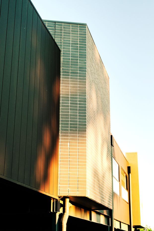 Upward-looking exterior architectural photograph of a contemporary multi-storey building. The facade combines dark vertical metal cladding with a prominent translucent or glazed grid-like wall and warm-toned panels, creating strong geometric planes and cantilevered forms. Bright low-angle sunlight highlights the facade against a pale sky.