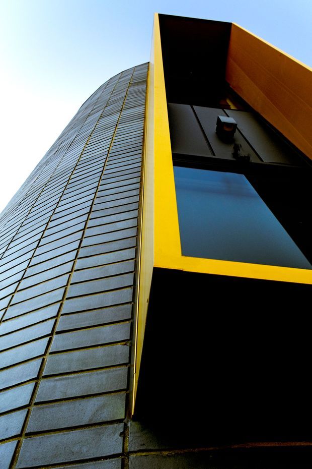 Low-angle view looking up at a tall contemporary building facade. The composition is dominated by a vertically rising glazed or translucent panel wall with strong horizontal divisions, contrasted by a bold yellow/orange projecting frame and deep black recessed glazing. The image emphasizes height, geometric lines, dramatic perspective, and an urban commercial or institutional facade against a pale blue sky.
