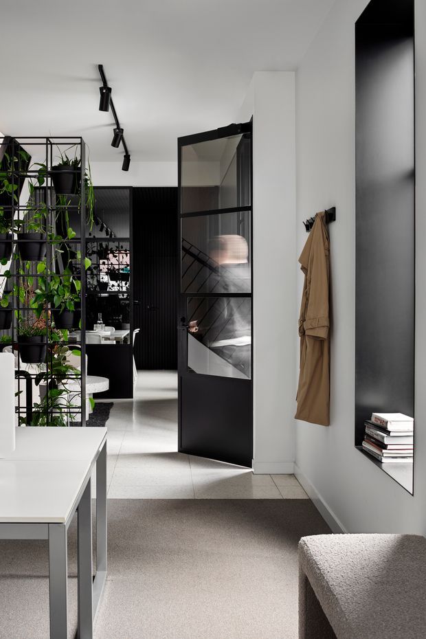 A bright, refined modern interior viewed from an entry or living area. The space features white walls, pale tiled flooring transitioning to a soft grey carpet, black-framed glazed internal doors, a black ribbed feature wall, a full-height mirror, minimalist black track lighting, and a tall black metal plant partition. A coat hangs on wall hooks, with a partial white table and upholstered bench in the foreground. Reflections through the glazed door reveal a stairway and additional interior spaces.