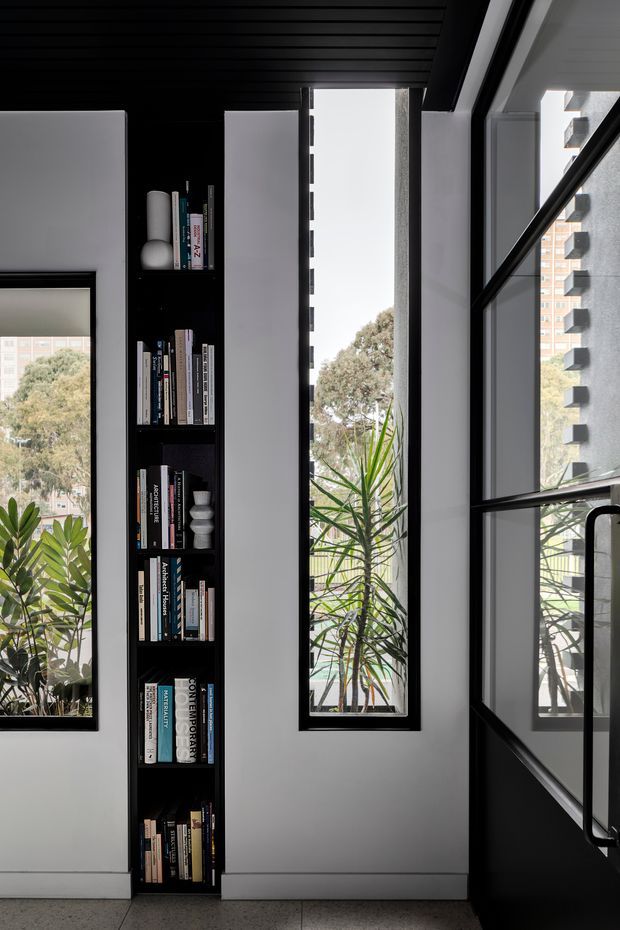 A narrow, vertically composed interior view featuring a built-in black bookcase filled with books and small decorative objects, set between white wall panels and tall slim windows. Black-framed glazing and a dark slatted ceiling create a strong monochrome architectural contrast, while indoor plants and views of neighbouring apartment buildings are visible through the windows. A black-framed glazed door or partition occupies the right side.