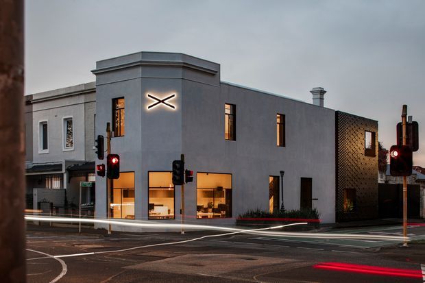 Street-level dusk view of a contemporary multi-storey commercial building on an urban corner. The building has a smooth dark grey rendered facade, large warm-lit ground-floor glazing, narrow upper-level windows, a projecting parapet, and a perforated brick or masonry side wall. Traffic signals, road markings, and neighbouring urban buildings frame the scene.