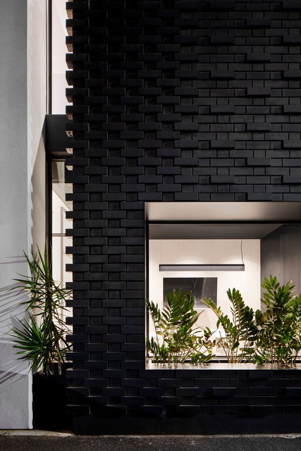 A close architectural view of a bold black brick facade forming a deep rectangular opening. The opening reveals a bright interior with indoor planting, a linear pendant light, artwork, and dark vertical wall detailing. A tall narrow window and additional greenery sit beside the projecting brickwork, creating a strong contrast between the dark textured masonry and light rendered wall.