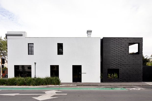 Street-facing view of a contemporary, flat-roofed building composed of a smooth white rendered volume and a contrasting dark brick-clad volume. The facade has sparse rectangular windows, a dark entry door, a projecting horizontal detail near the roofline, and a large square opening in the brick section. Low ornamental planting lines the pavement, with a marked urban street in the foreground.