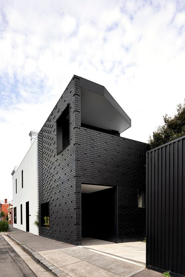 Street-level exterior view of a narrow contemporary multi-storey residence. The building has a strongly defined black brick facade, a projecting flat roof or upper-level canopy, minimalist dark-framed openings, and a contrasting white side volume. A tall black vertical-slat fence and concrete driveway line the frontage.