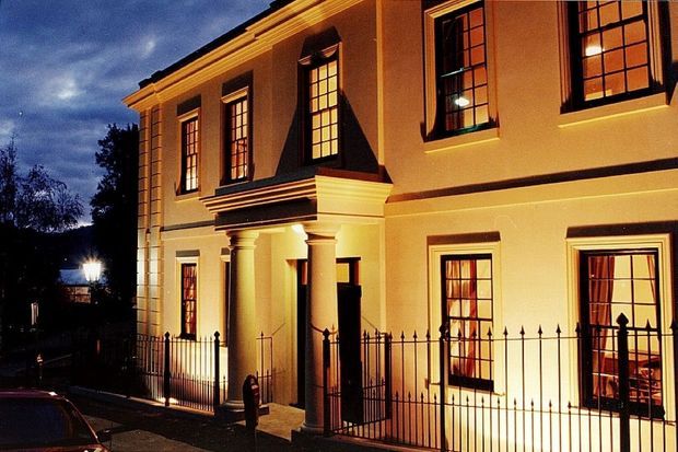 A warmly illuminated two-storey classical facade photographed at dusk. The cream-rendered building features strong symmetrical proportions, tall multi-pane sash windows with dark trim, a projecting central entrance portico with columns and entablature, decorative cornices, and a black wrought-iron front fence and gate.