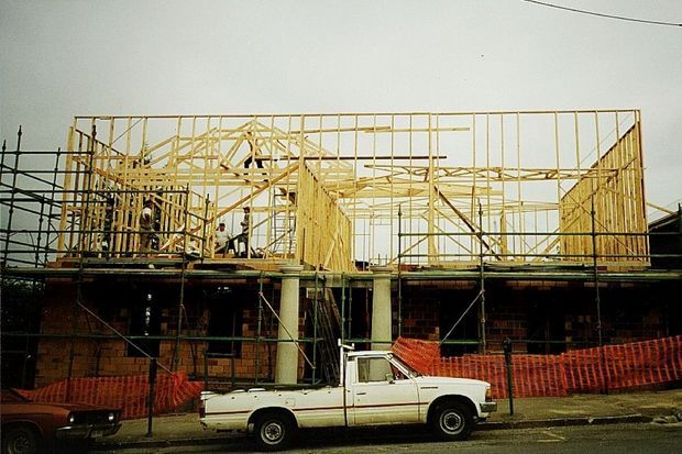 A partially constructed multi-storey residential building viewed from the street, with exposed timber wall and roof framing above brickwork, extensive scaffolding, temporary safety fencing, and a parked utility vehicle in the foreground.