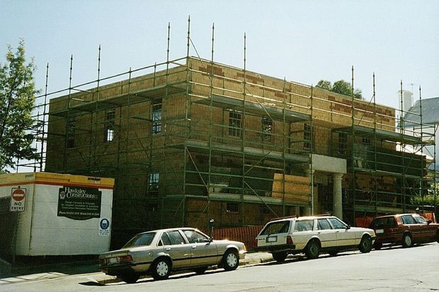 Street-level exterior photograph of a multi-storey brick building under construction, enclosed in extensive metal scaffolding. Temporary site fencing and a construction trailer are visible in the foreground, with parked cars along the street.
