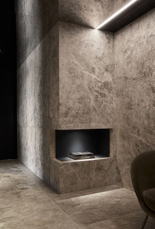 A moody, minimalist interior featuring a monolithic floor-to-ceiling fireplace or architectural feature wall clad in large-format veined stone-look slabs. A recessed black niche with subtle lighting displays books, while linear concealed lighting and a dark slatted ceiling detail add drama. Matching stone-look flooring and a partial rounded upholstered chair are visible.