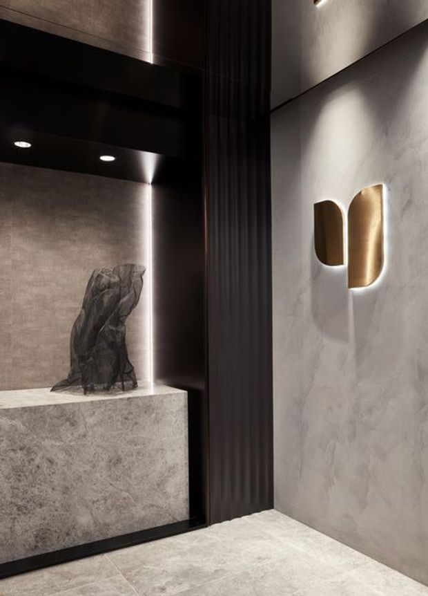 A dramatic, refined interior architectural detail featuring a grey stone-clad pedestal, dark ribbed vertical partition, black reflective wall and ceiling surfaces, polished grey flooring, and a sculptural wall sconce with a warm brass-gold finish. Concealed linear lighting and recessed downlights create a moody gallery-like atmosphere.