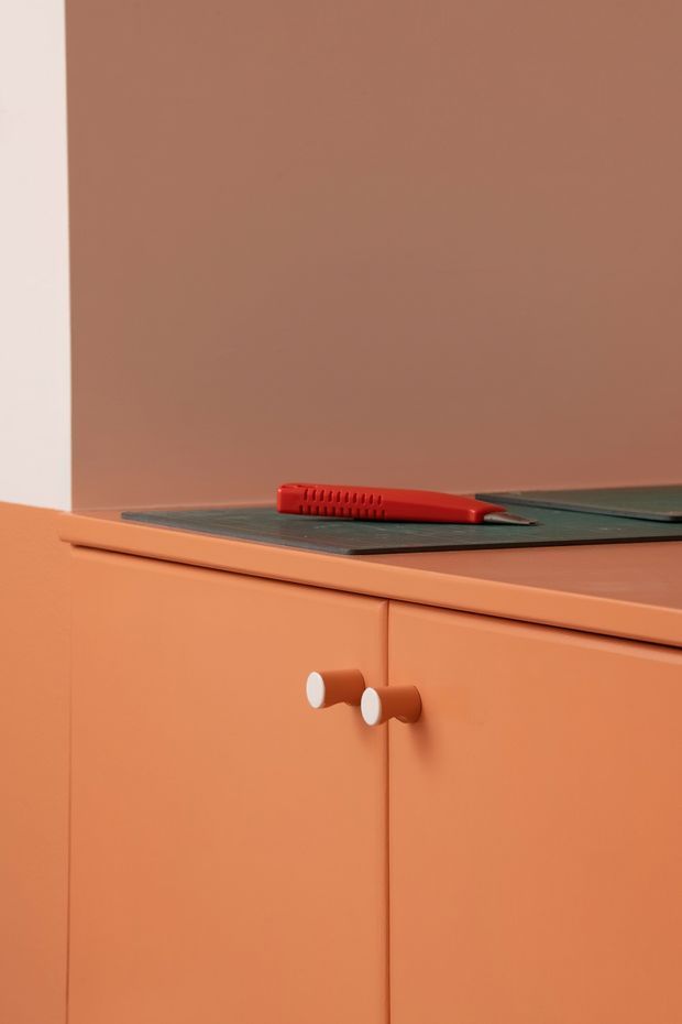 Close-up detail of a monochromatic orange kitchen cabinet and benchtop, featuring flat-panel cabinetry with cylindrical orange knobs and a dark flush-mounted cooktop. A red kitchen utensil rests on the cooktop, while a pale vertical wall edge provides contrast.