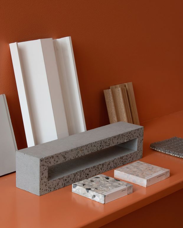 A styled materials display featuring a grey terrazzo-like concrete block with a recessed shelf, white and timber sample boards leaning against a terracotta wall, and several terrazzo or stone sample tiles arranged on a matching terracotta surface. The image is a close-up product/material composition rather than a complete room.