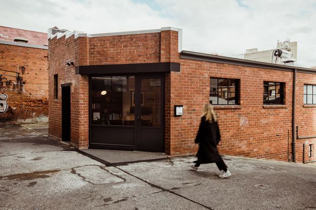 Exterior view of a low-rise red brick commercial or industrial-style building, featuring a recessed dark-framed glazed entrance, black-framed windows, flat rooflines, and a paved urban service lane in front. The exposed brickwork and utilitarian facade dominate the image.