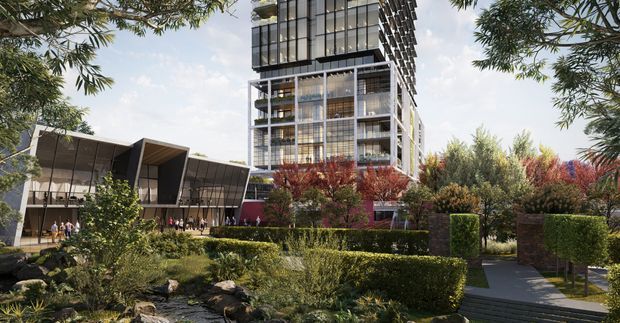 A landscaped urban development viewed from a garden walkway, featuring a tall contemporary multi-storey building with extensive glass curtain walling, projecting balconies and dark structural framing. A lower pavilion with angular roof forms sits to the left, surrounded by clipped hedges, ornamental planting, trees and a small rocky garden stream or water feature.