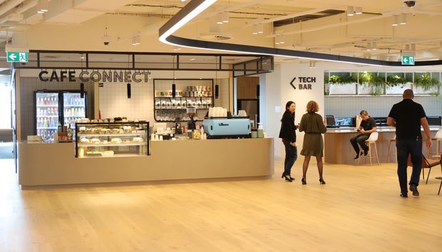 A bright contemporary commercial café and tech bar interior in an open-plan workplace or public venue. The café counter includes refrigerated beverage displays, a pastry display case, shelving, and a turquoise espresso machine, with signage reading “CAFE CONNECT” and “TECH BAR.” Pale timber flooring, a beige service counter, black-framed glass partitions, exposed services, hanging greenery, and linear modern lighting define the space. Several people are present, with seating and a secondary counter visible to the right.
