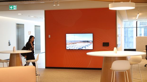 Contemporary commercial office breakout and collaboration area with high round tables, stools, lounge seating, pendant lighting, and a wall-mounted display. A bold orange-red feature wall with subtle contour-line graphics forms the focal point, while glazed windows and an open circulation area create a bright workplace setting.