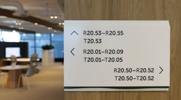 A close-up of a white directional wayfinding sign mounted on a vertical wood-panelled wall. The sign uses black text and simple arrow symbols to indicate room ranges, while a softly blurred contemporary office or commercial interior with tables, chairs, pendant lighting, and glazing is visible in the background.