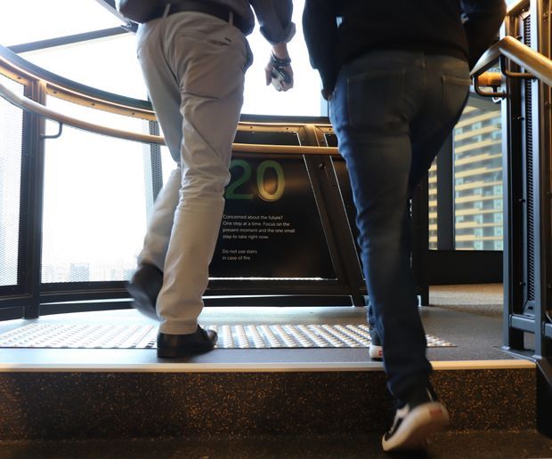 A low-angle view of two people ascending illuminated steps inside a modern lift or stair access area. The space features dark metal framing, perforated metal flooring, curved timber-toned handrails, glass panels, and bright daylight through surrounding glazing. A large green “20” and safety instructions are visible on the central panel, with a high-rise building seen outside.