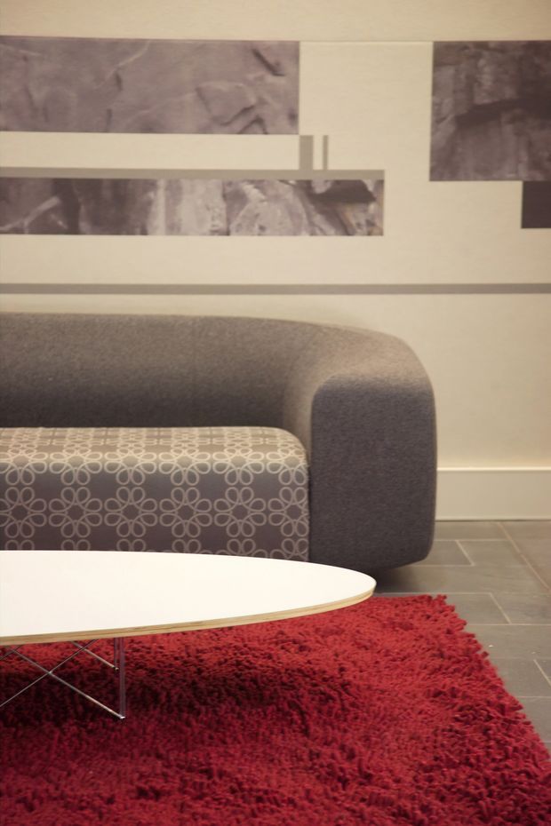 A close-up contemporary living room vignette featuring a rounded grey upholstered sofa, a patterned ottoman or lounge cushion, a low white oval coffee table with slender metal legs, and a deep red shag rug. The wall has abstract grey artwork or panels, while the floor is finished in large grey rectangular tiles. The palette is muted grey and cream with a strong red accent, creating a stylish mid-century-inspired lounge setting.