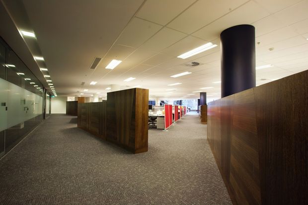 A modern open-plan commercial office interior viewed from a circulation area. The space has low acoustic ceiling tiles with recessed fluorescent lighting, grey carpet flooring, dark timber-clad partition walls and cabinetry, a glazed office wall on the left, and colourful workstation partitions visible in the central background. A large dark cylindrical structural column provides a prominent feature.