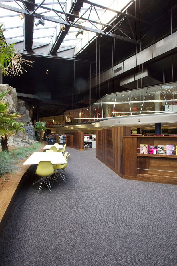A large contemporary commercial interior with an open-plan workspace or hospitality-style lounge. The space features dark exposed steel roof trusses, extensive skylights and glazing, suspended linear lighting, a mezzanine-like upper level, dark timber built-in counters and shelving, grey carpet, and a dramatic indoor rock and tropical planting feature. Lime-green and white dining chairs surround small tables in the foreground.