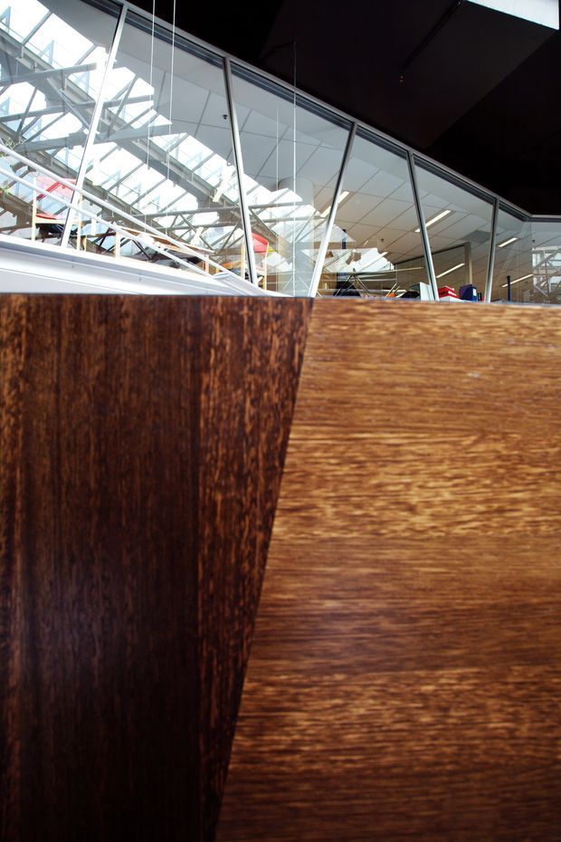 Interior architectural detail showing a dark timber-clad partition or built-in wall in the foreground, with full-height glazed partitions behind it. The glazing reveals a bright contemporary commercial or institutional interior with exposed steel roof structure, suspended linear lighting, white ceiling panels, and glimpses of colorful furnishings. The image emphasizes timber texture, glass, structure, and daylight rather than a specific room function.