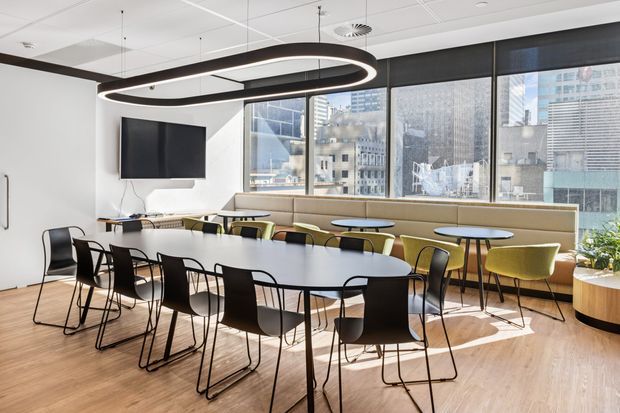 Bright contemporary commercial break-out or meeting area with a long communal dining table, black and olive-green chairs, built-in upholstered banquette seating, a wall-mounted television, and large windows overlooking a dense urban setting. The space has light timber flooring, white walls, a suspended acoustic ceiling, and a distinctive oversized oval pendant light.