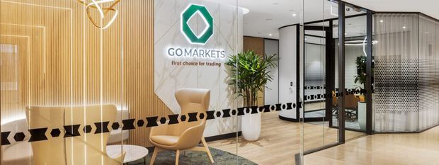 A polished commercial office reception and waiting area with a branded GOMARKETS feature wall, vertical timber-look slatted wall treatment, glass-partitioned meeting or office rooms, pale timber flooring, contemporary lounge seating, indoor plants, and black geometric privacy decals across the glazing.