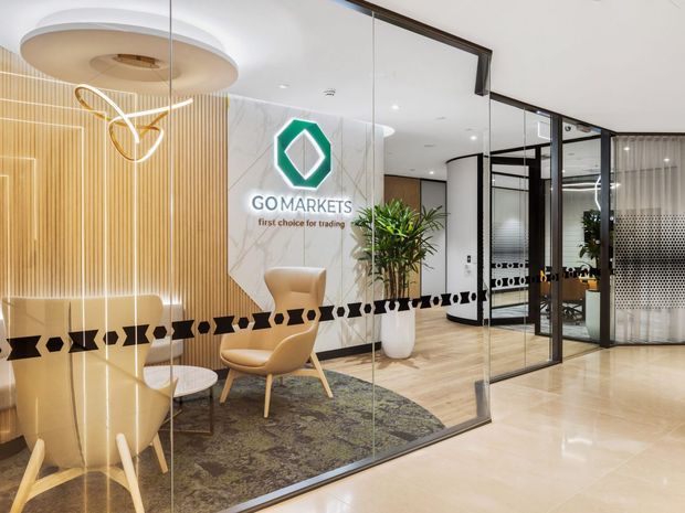 A polished commercial office reception and waiting area viewed through full-height glass partitions. The space features a curved upholstered lounge chair, small round table, patterned rug, indoor planting, a wood-slat feature wall, marble-look branded wall panel, black-framed glazing, and decorative pendant lighting. The GoMarkets logo is prominently displayed, with additional glazed meeting or office areas visible beyond.