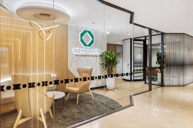 A polished corporate office reception and waiting area viewed through glass partitions. The space features warm vertical timber wall battens, a marble-look feature wall with the GOMarkets logo, a sculptural circular ceiling light, contemporary lounge chairs and side tables, indoor planting, pale timber flooring, and black-framed glazed meeting-room partitions with patterned privacy film.