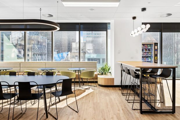 A bright contemporary commercial break-out and dining area with extensive floor-to-ceiling city-facing glazing, light timber flooring, communal dining tables, varied modern chairs, a high-top table, and distinctive pendant lighting. A compact vending and refreshment area is visible at the rear.
