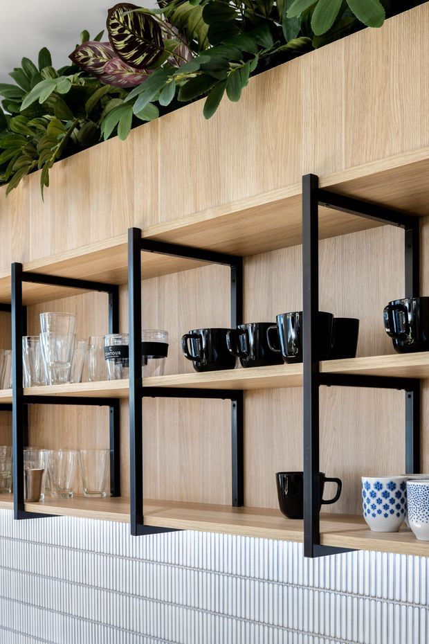 Close-up of a contemporary built-in shelving and display area, featuring pale timber shelving with black metal supports, glassware and ceramic mugs, a vertical white ribbed tile wall below, and lush greenery arranged along the top.