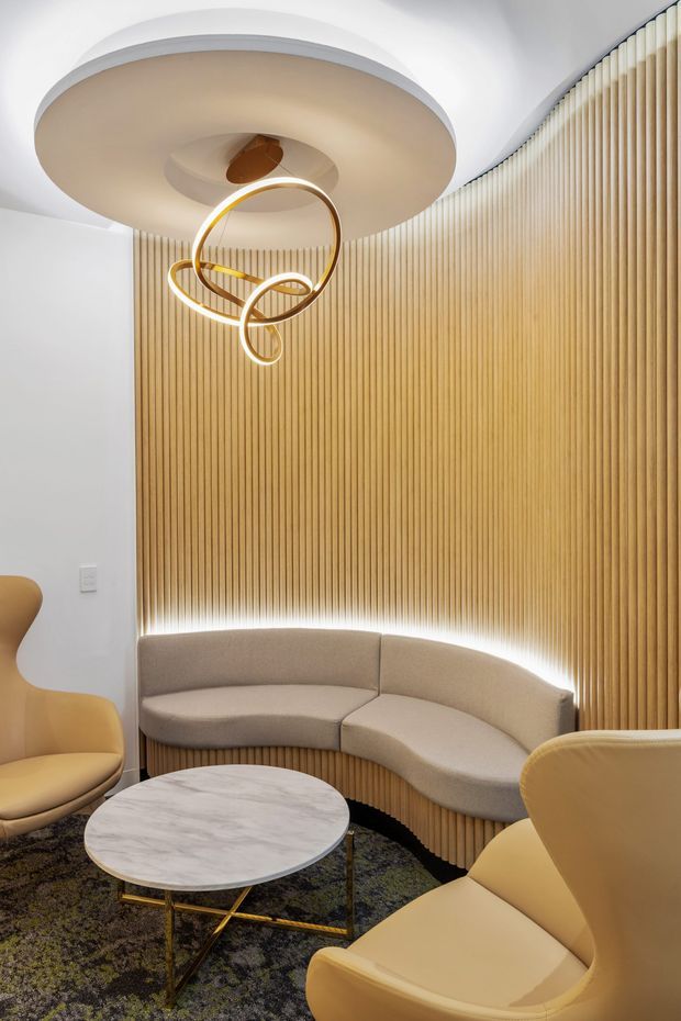 A refined contemporary lounge or reception area with a curved upholstered banquette wrapping around a ribbed timber feature wall. Warm integrated lighting glows beneath the seating and along the ceiling, complemented by sculptural gold pendant lighting, rounded mustard armchairs, and a circular marble-top coffee table. The space has a softly curved, high-end hospitality feel with layered neutral and warm wood tones.