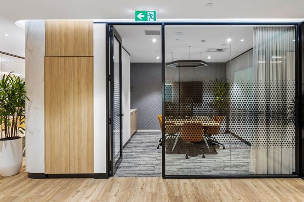 Modern commercial office interior showing a glazed meeting room with black-framed glass walls, dotted privacy film, a conference table and orange chairs. The room is accessed from a light timber-finished corridor, with integrated wood wall panelling, patterned carpet, curtains and contemporary ceiling lighting.