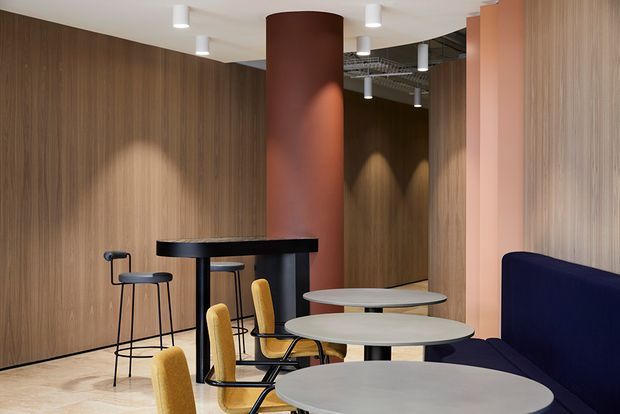A contemporary hospitality or commercial lounge and dining area with curved wood-paneled walls, a prominent rust-coloured cylindrical column, round café tables, mustard upholstered chairs, black bar-height seating, and a dark navy banquette. Recessed and pendant-style ceiling lights create a refined, warm atmosphere.