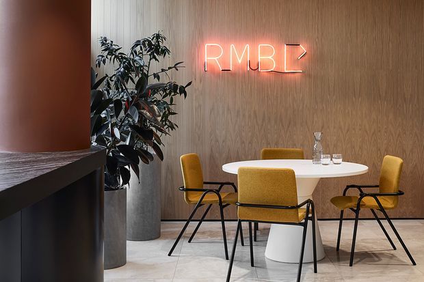 A stylish contemporary commercial dining or meeting area with a round white table, mustard upholstered chairs, black metal frames, a light wood-panelled feature wall, pale tiled flooring, a tall indoor plant, and a glowing neon “RMB” sign. A curved dark reception or counter element occupies the left foreground.
