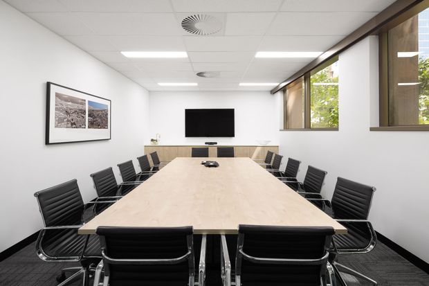 A bright, contemporary conference room with a long pale timber meeting table surrounded by black leather office chairs. The room has white walls, a suspended acoustic ceiling with recessed lighting, dark carpet flooring, large windows along one side, framed wall photography, and a wall-mounted display screen.
