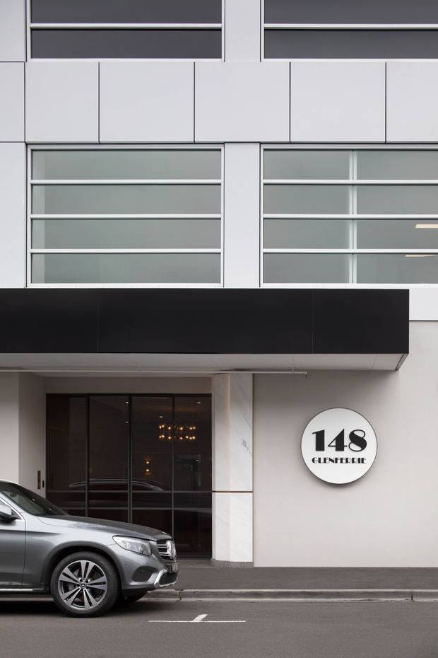 Street-level view of a contemporary multi-storey commercial building facade. The entrance is framed by dark glazed doors, a projecting black canopy, pale stone or concrete wall surfaces, and large horizontal strip windows with light-coloured frames. A circular sign marks the building as 148 Glenferrie. A parked car is visible in the foreground.