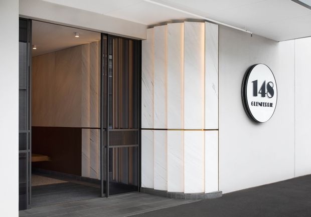 A contemporary commercial building entrance with a recessed lift or lobby opening behind dark metal-framed glazed and slatted doors. The facade features tall white marble-look wall panels with subtle veining, dark grey tiled paving, and a circular wall sign marked “148”. The space is clean, refined, and predominantly monochromatic with warm timber visible inside the lobby.