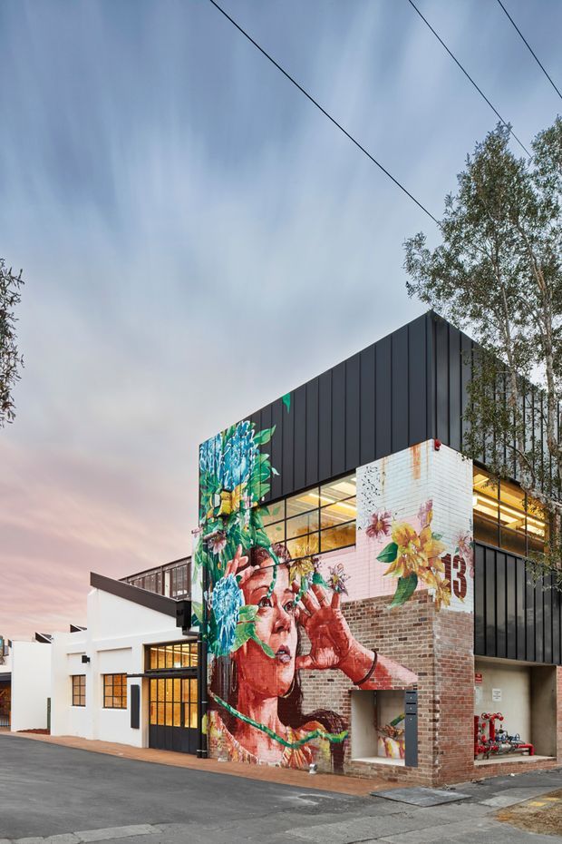 Exterior view of a contemporary multi-storey commercial or industrial building at dusk. The building combines black vertical metal cladding, exposed brickwork, large glazed sections, and white rendered volumes. A large colourful floral and portrait mural dominates the street-facing facade, with visible exterior services and an asphalt forecourt.